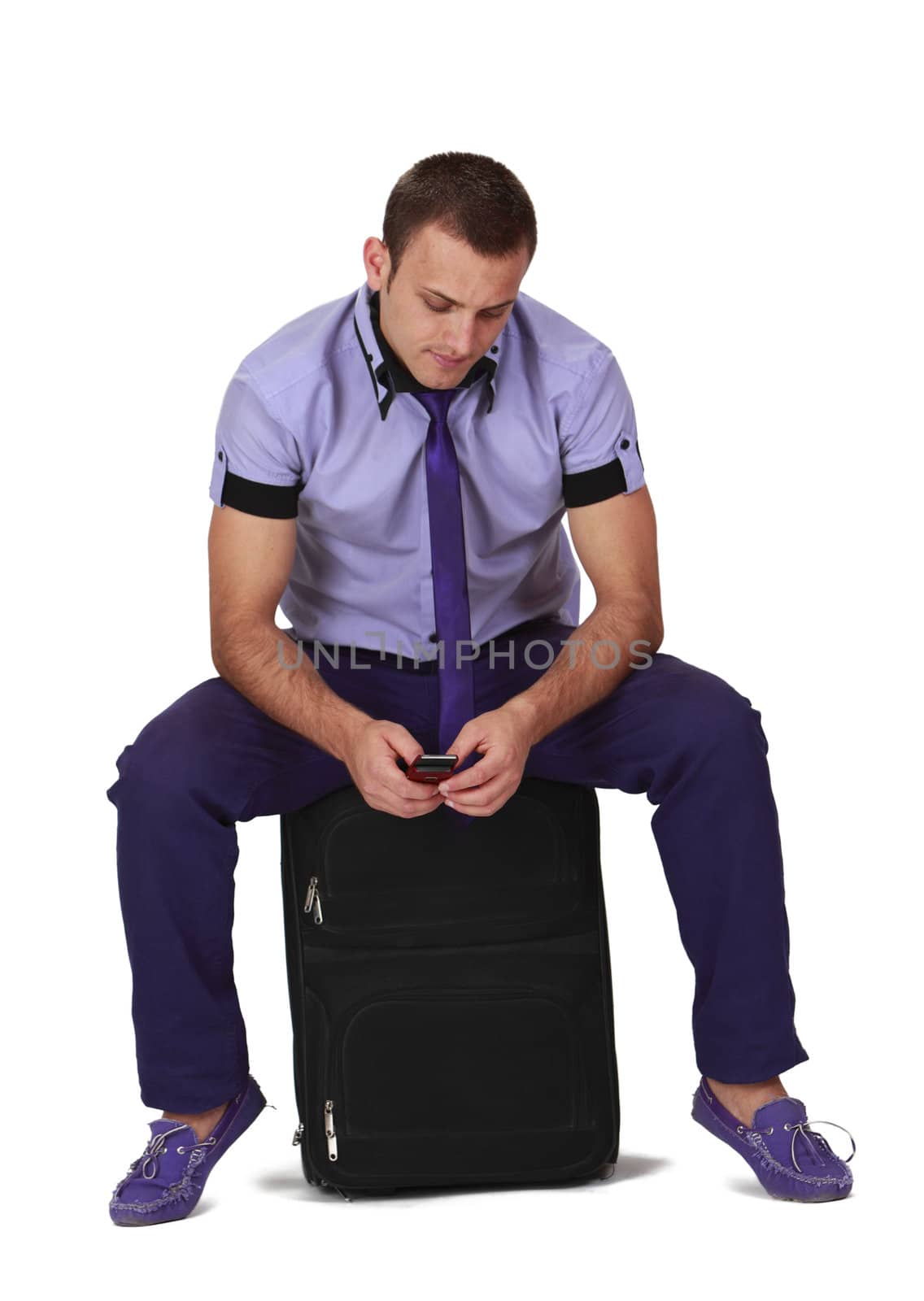 Young man sitting on a suitcase and cecking his mobile phone, isolated against a white background.