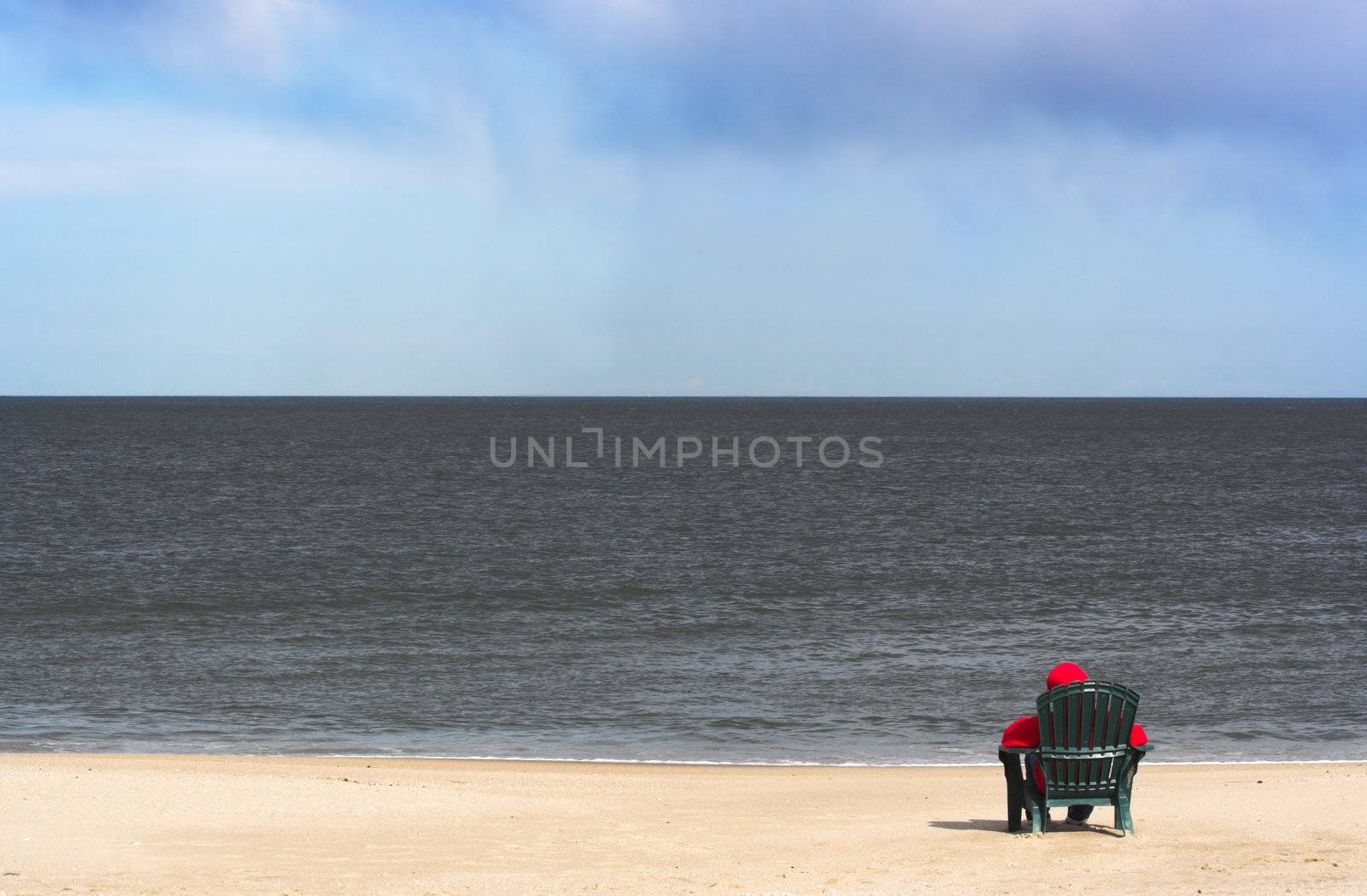 A young person in a hooded red sweatshirt sitting alone on a chair in the sand at the beach. The hood of the sweatshirt is on indicating it is a chilly day. The person is looking out over the ocean.