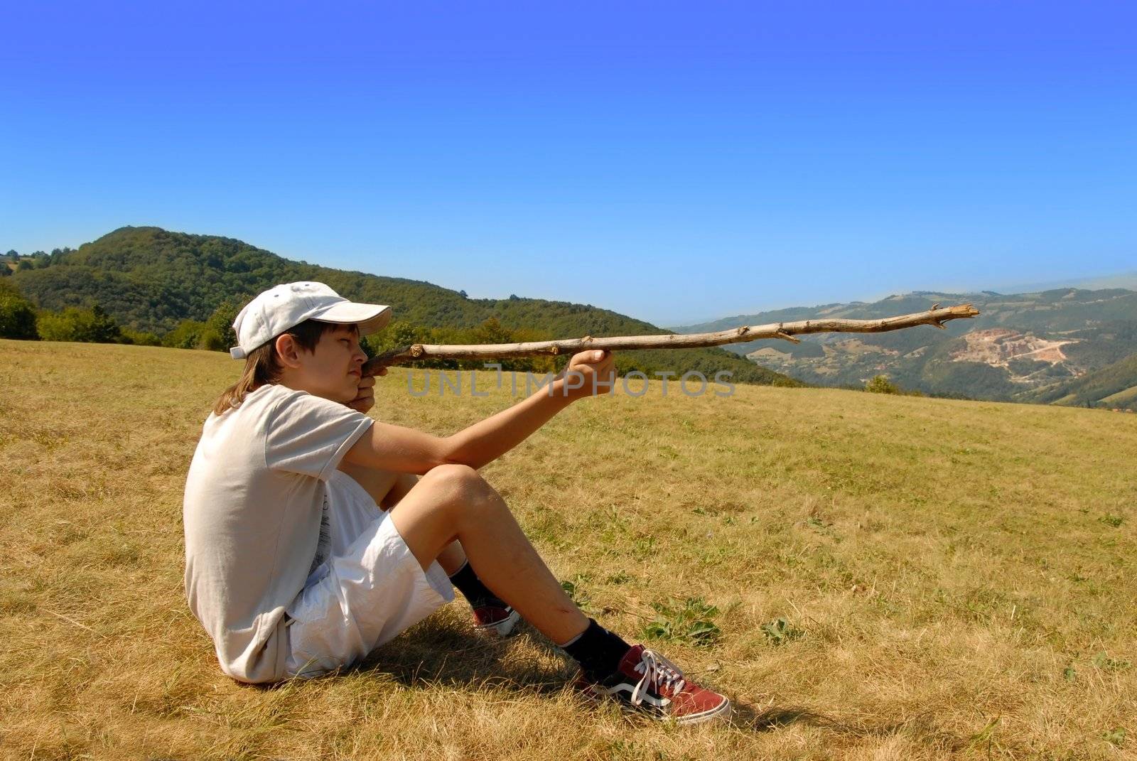 boy sitting on mountain meadow aiming with a stick as a gun