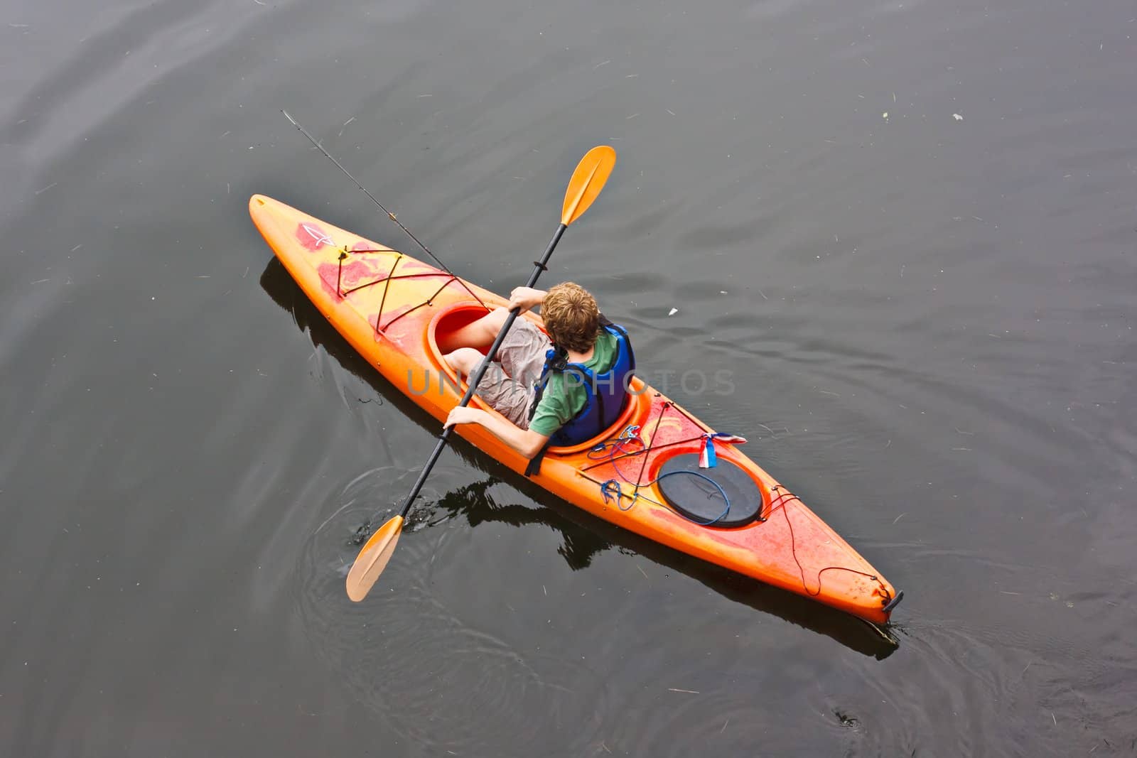A teenage boy paddling a kayak on a lake. There is a fishing pole in the kayak with him.