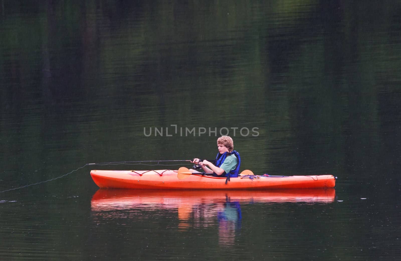 A young teenage boy fishing from a kayak in the evening. It is raining and streaks of rain can be seen against the lake background.