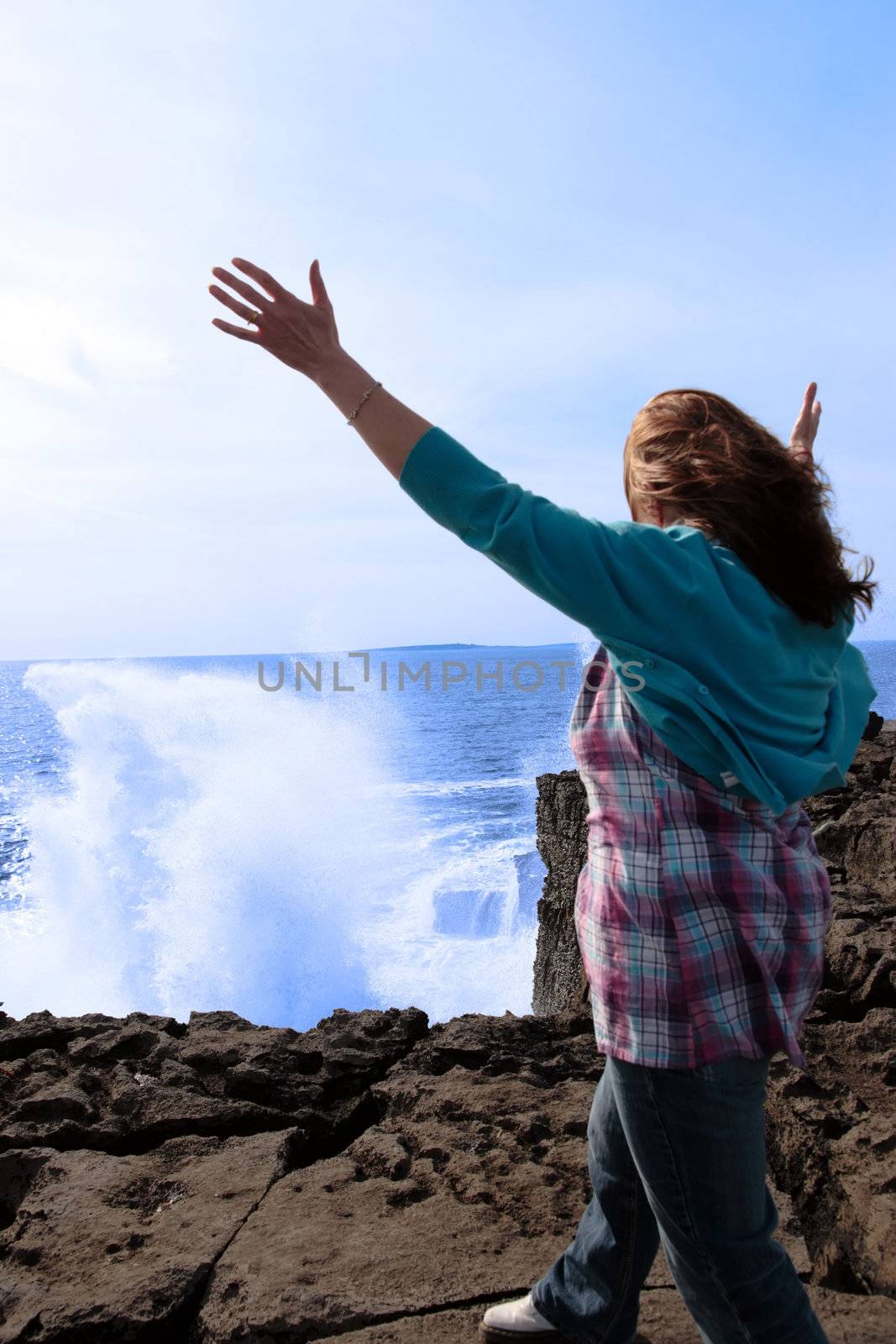 a lone woman raising her arms in awe at the powerful waves on the cliffs edge in county clare ireland