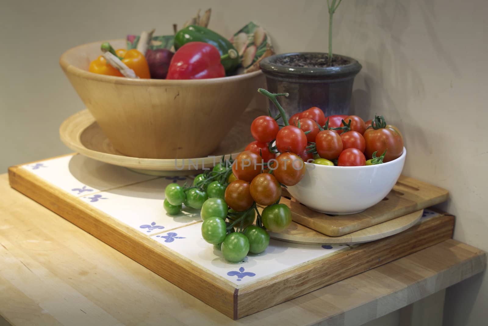 A kitchen setting with a white ceramic bowl of freshly picked small green, red and yellow coloured tomatos cascading from the bowl, set on a wooden kitchen island unit. A bowl of colored peppers is set to the back of the image.