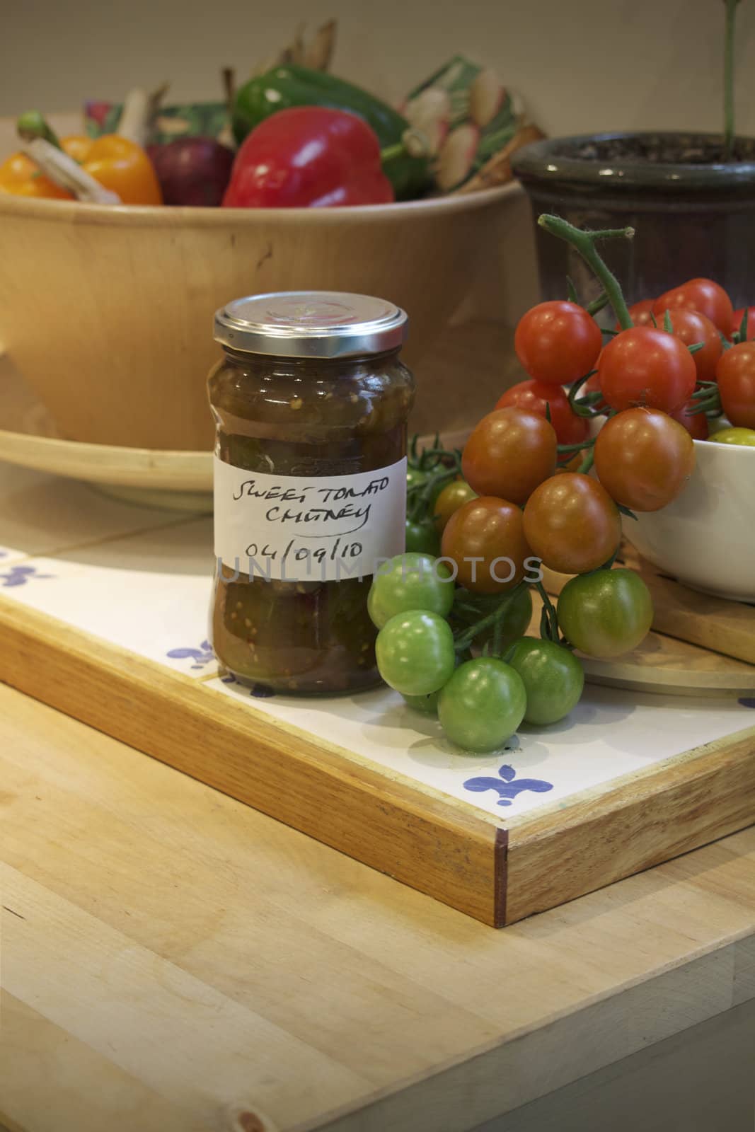 A kitchen setting with a white ceramic bowl of freshly picked small green, red and yellow coloured tomatos cascading from the bowl with a jar of homemade sweet tomato chutney in front with a handwritten label. Set on a wooden kitchen island unit. A bowl of colored peppers is set to the back of the image.