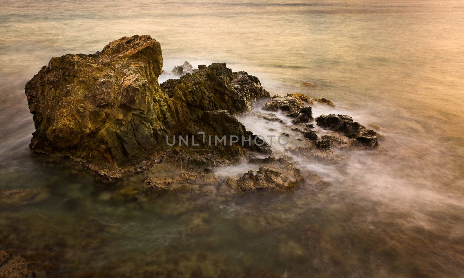 Slow exposure blurs the waves in the ocean as they break over a rocky outcrop at sunset