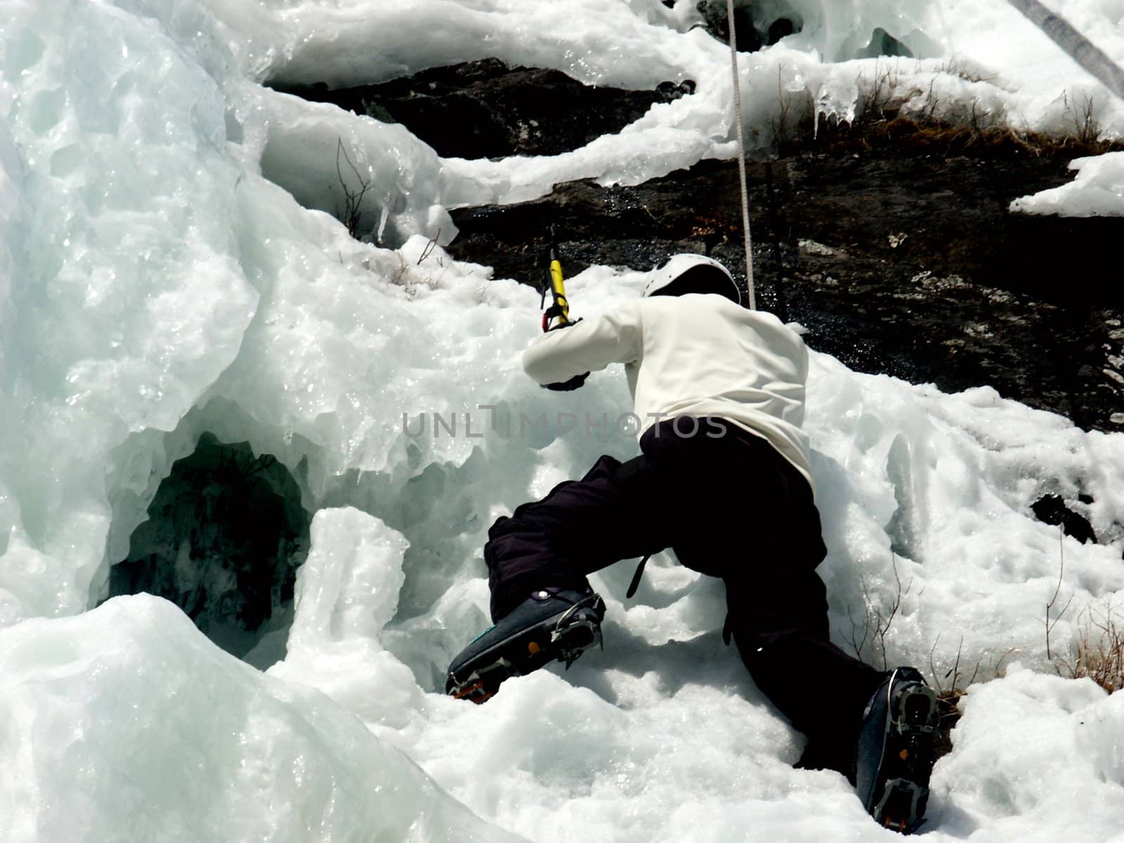 A man climbs up a frozen waterfall using ice axes (tools) and crampons, on a top-rope belay system.