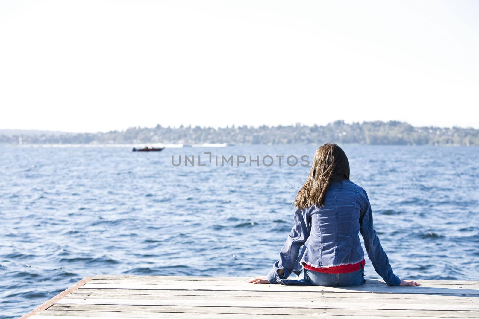 Girl sitting alone on dock by lake, looking out over water.