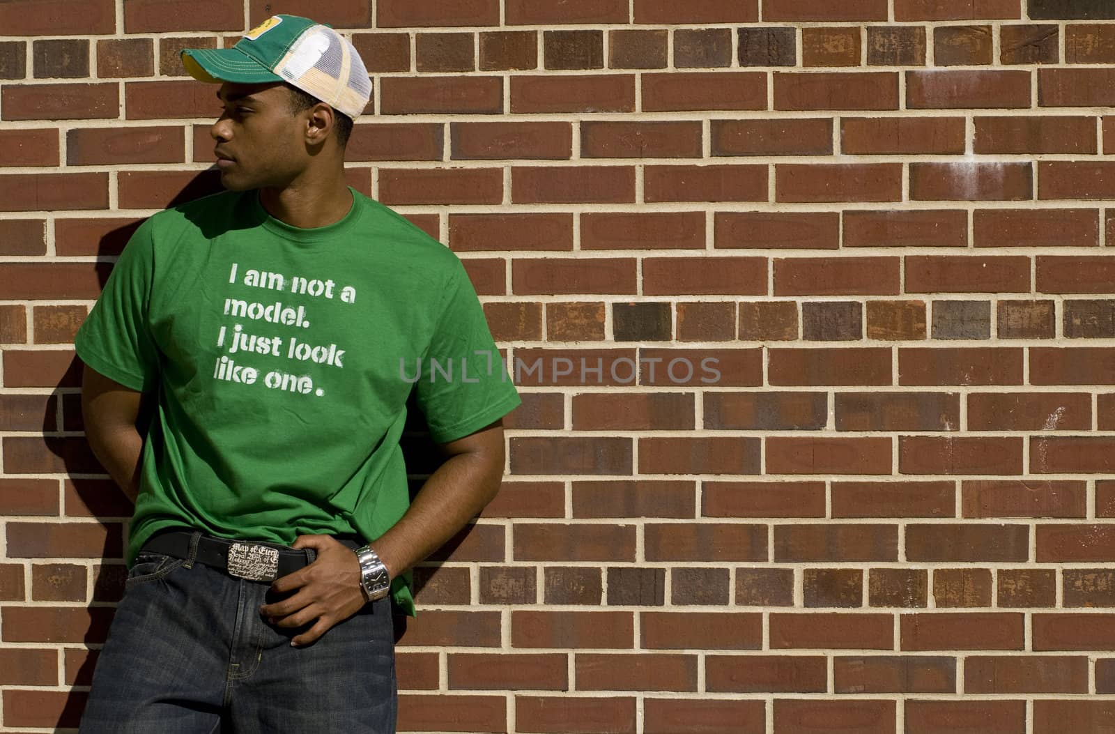Attractive young African American male playing posing in a green t-shirt and jeans against a brick wall.