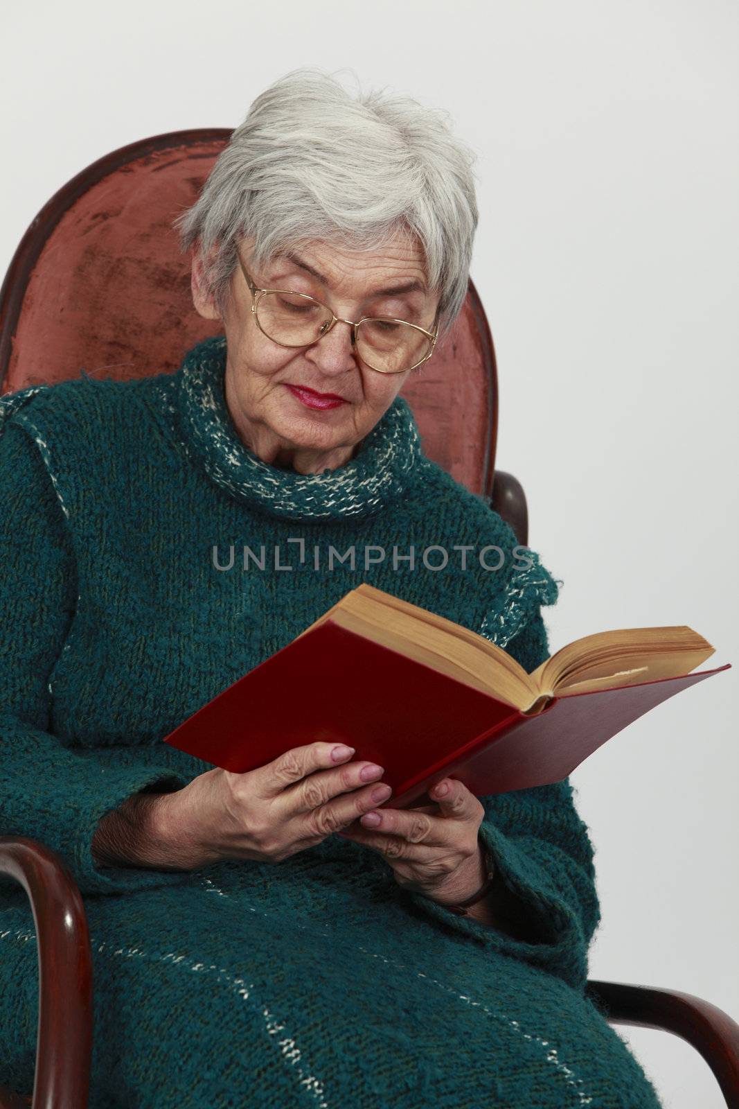 Portrait of an old woman reading a red book against a gray background.