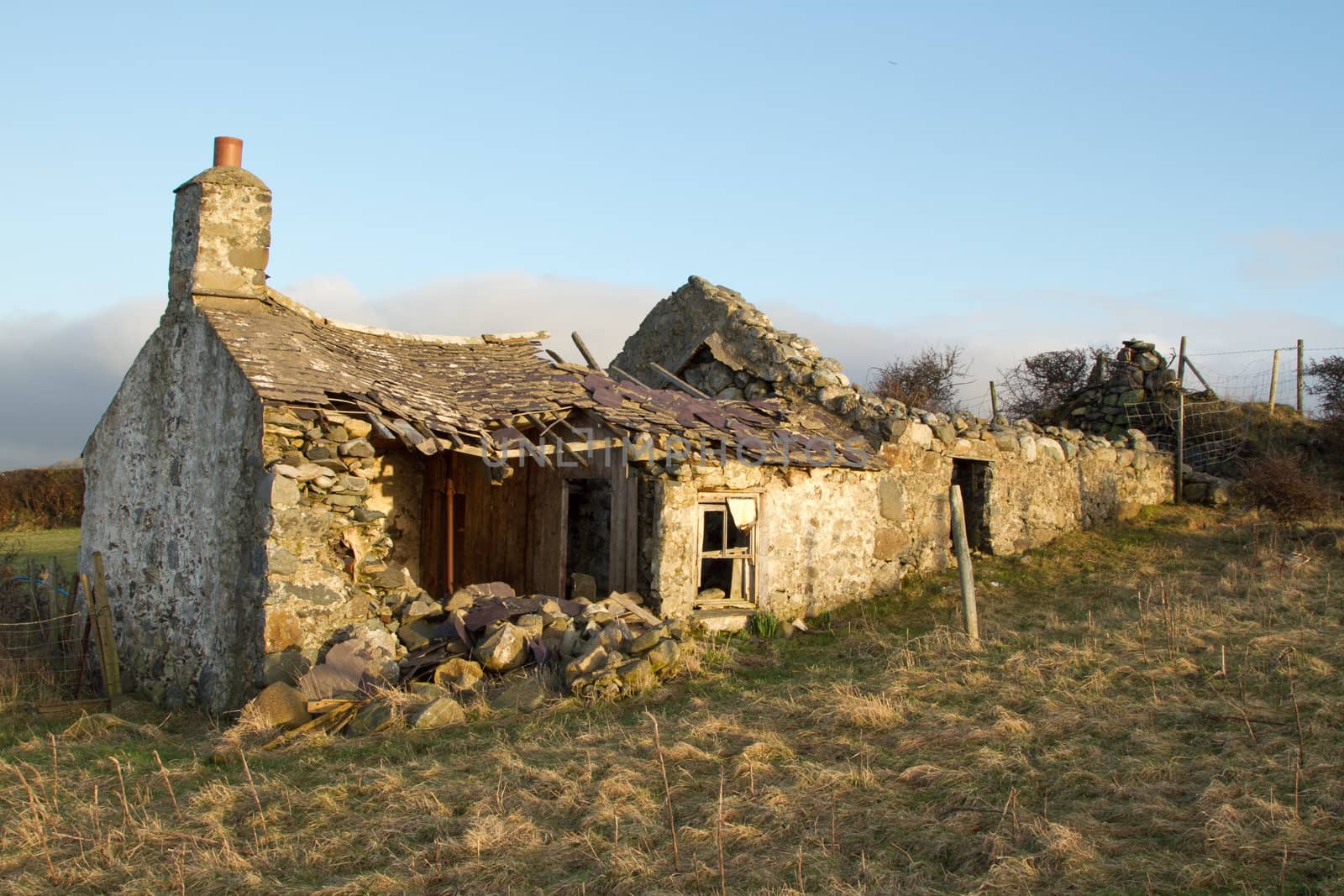 Derelict abandoned building, cottage, with crumbling walls and roof on grass with a blue sky.