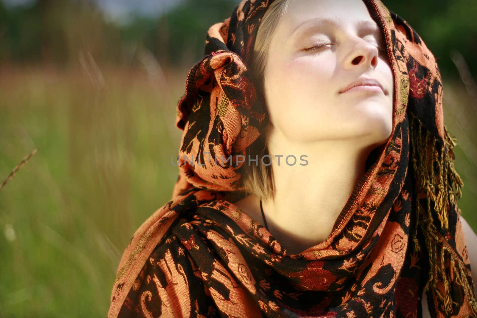 Young woman with traditional kerchief over head sitting in meadow and enjoying summer wind with closed eyes.