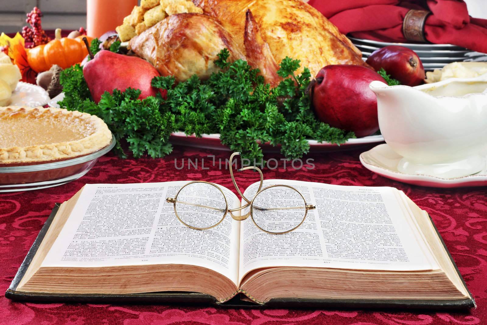 Open Bible with glasses lying on a holiday dinner table with prepared turkey and fixings in background.