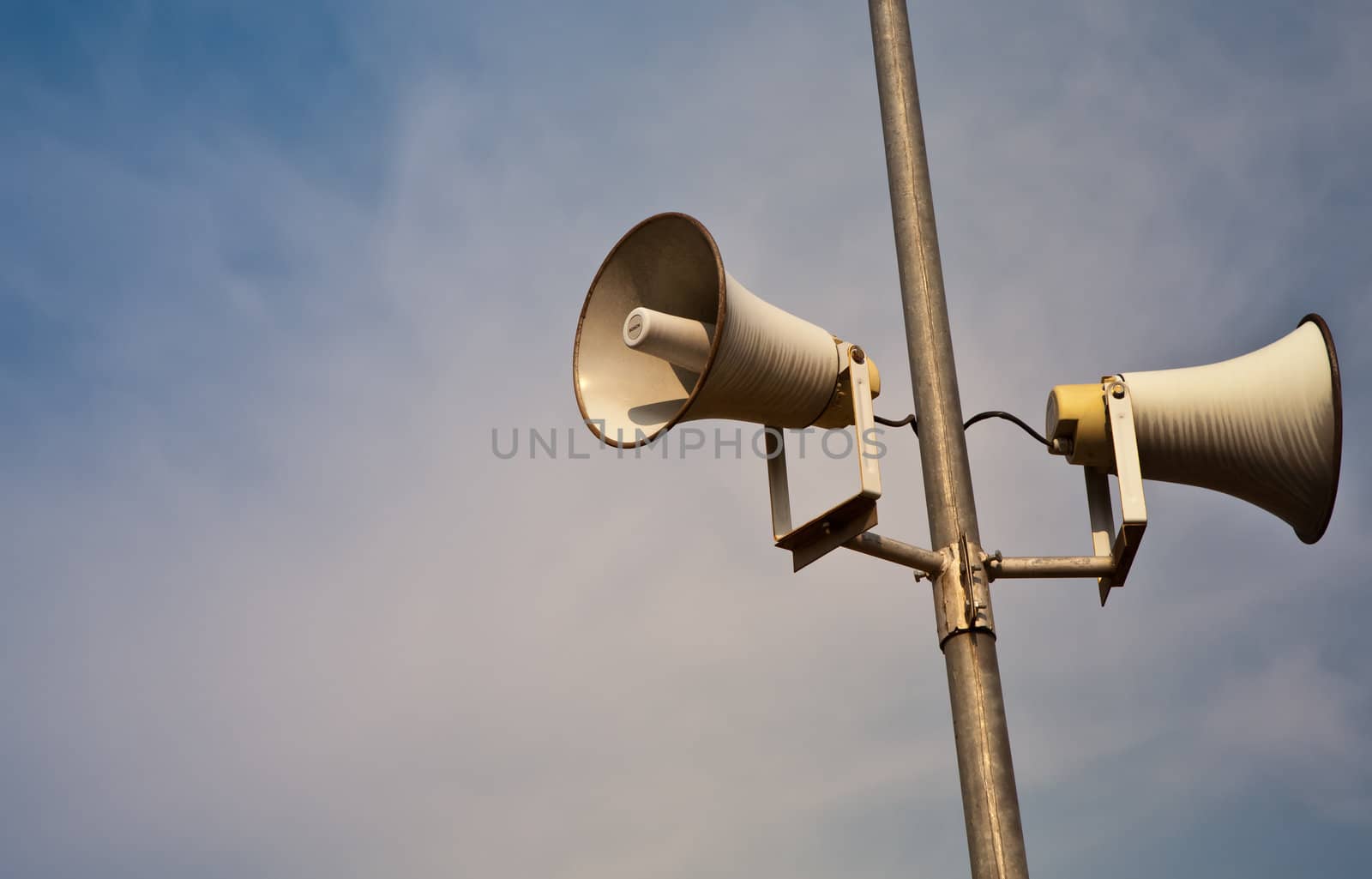 Public address horn loud speakers on metal pole over blue sky.