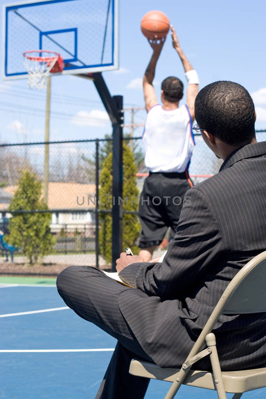 A basketball coach in a business suit observing a player on the team.   He could be also be recruiter trying to scout him.