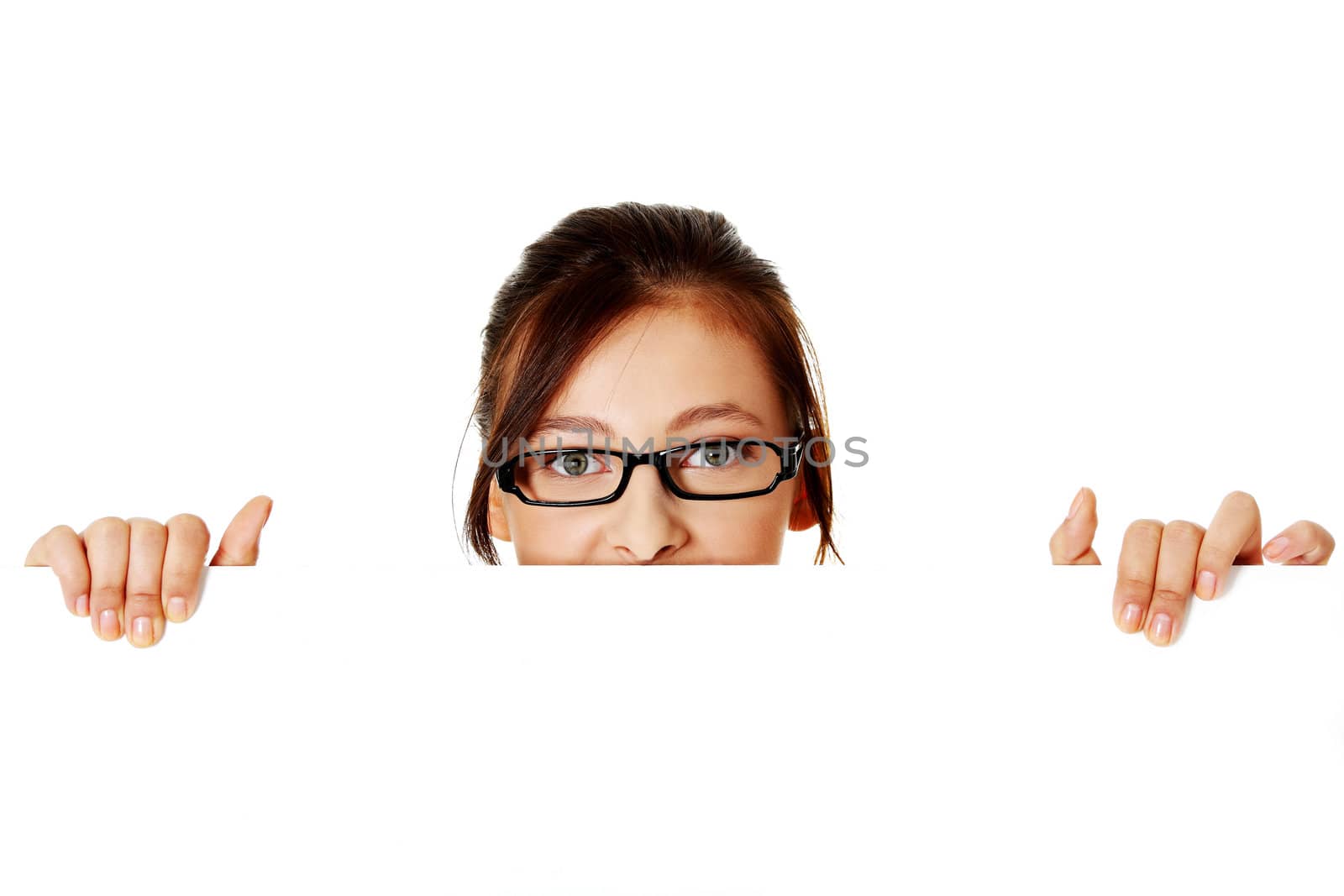 Young pretty girl in glasses hiding behind sheet of paper over white background.