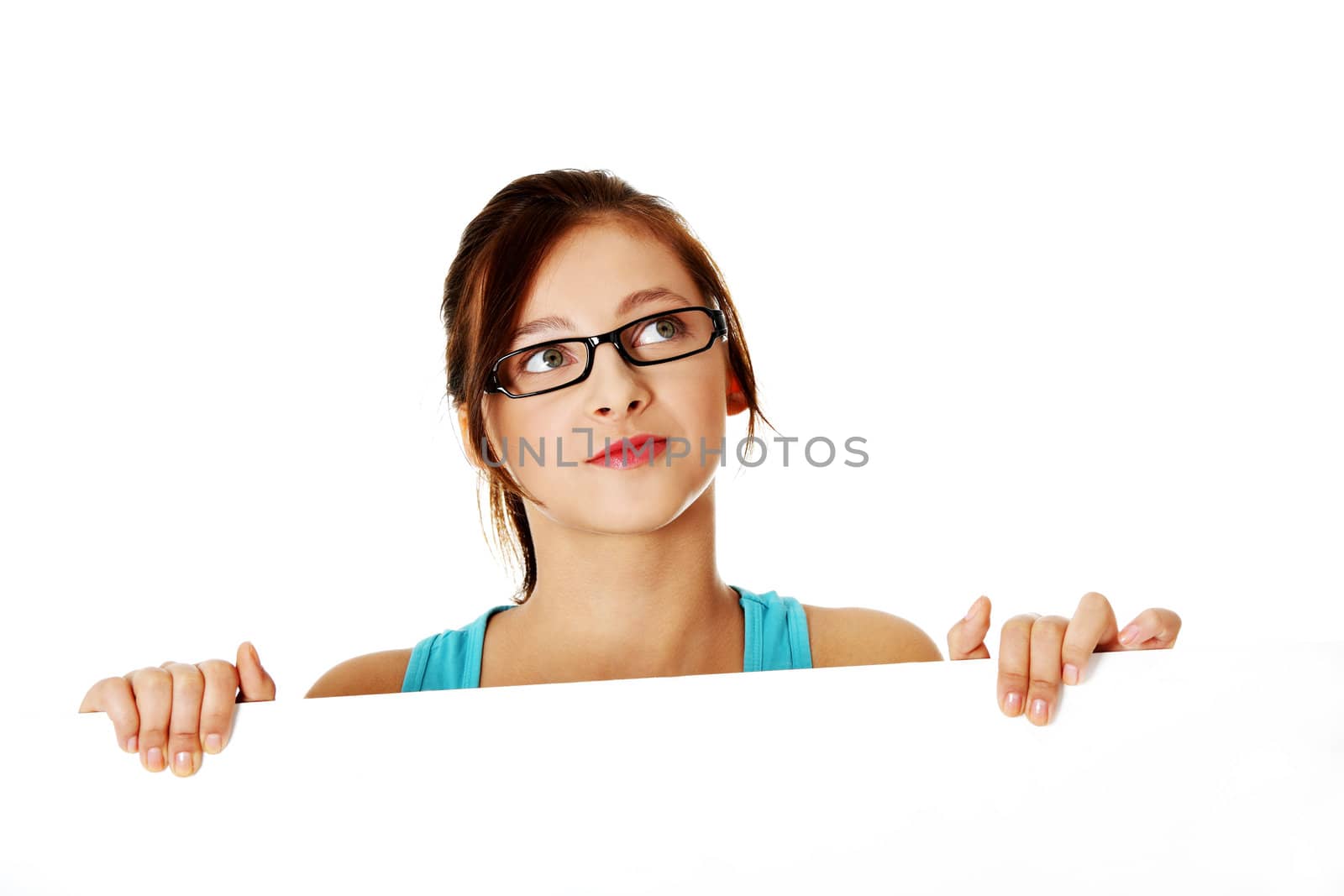 Young thinking girl in glasses hiding behind sheet of paper over white background.