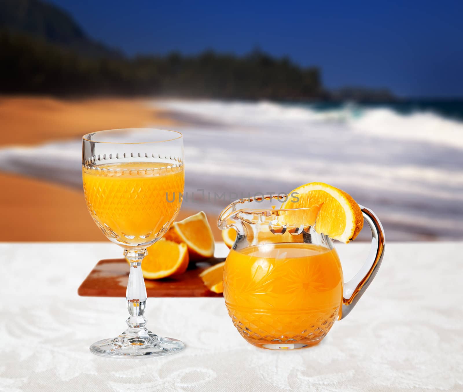 Orange juice in cut glass goblet with small jug and slices of orange in front of sandy beach and set on white linen tablecloth