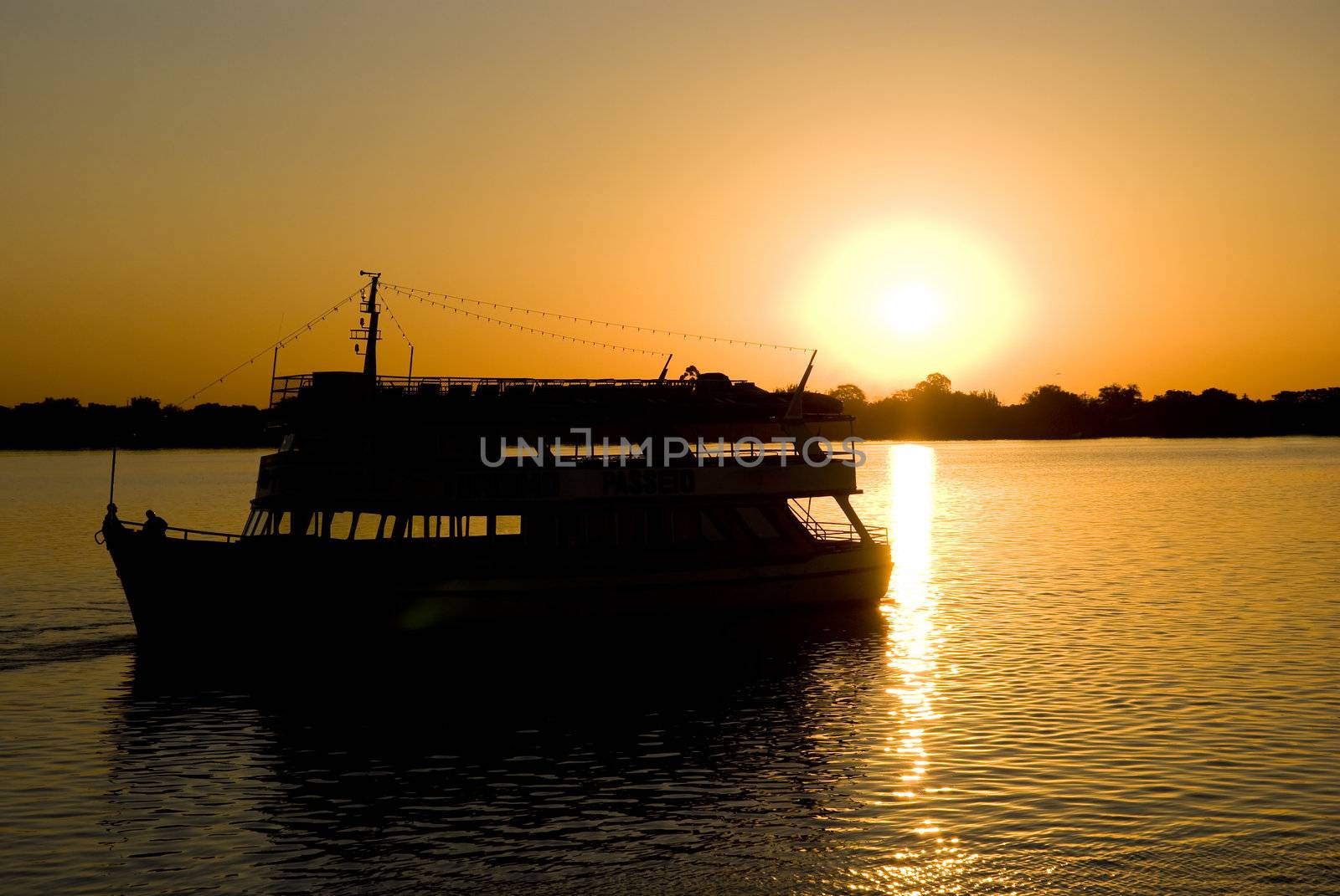 Silhouette of a small boat sailing away from setting sun, with deep orange hue.