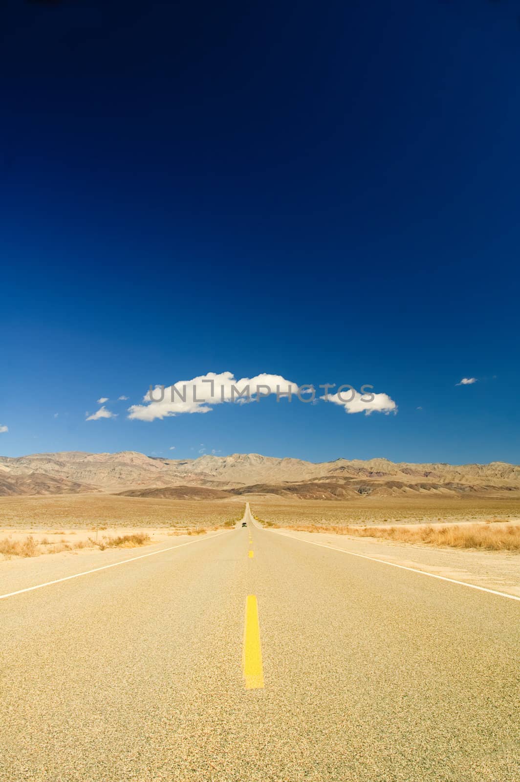 long straight road across Death Valley with a distant motor vehicvle coming towards the camera, a deep blue sky late in the afternoon, and a small puffy clouds in the sky