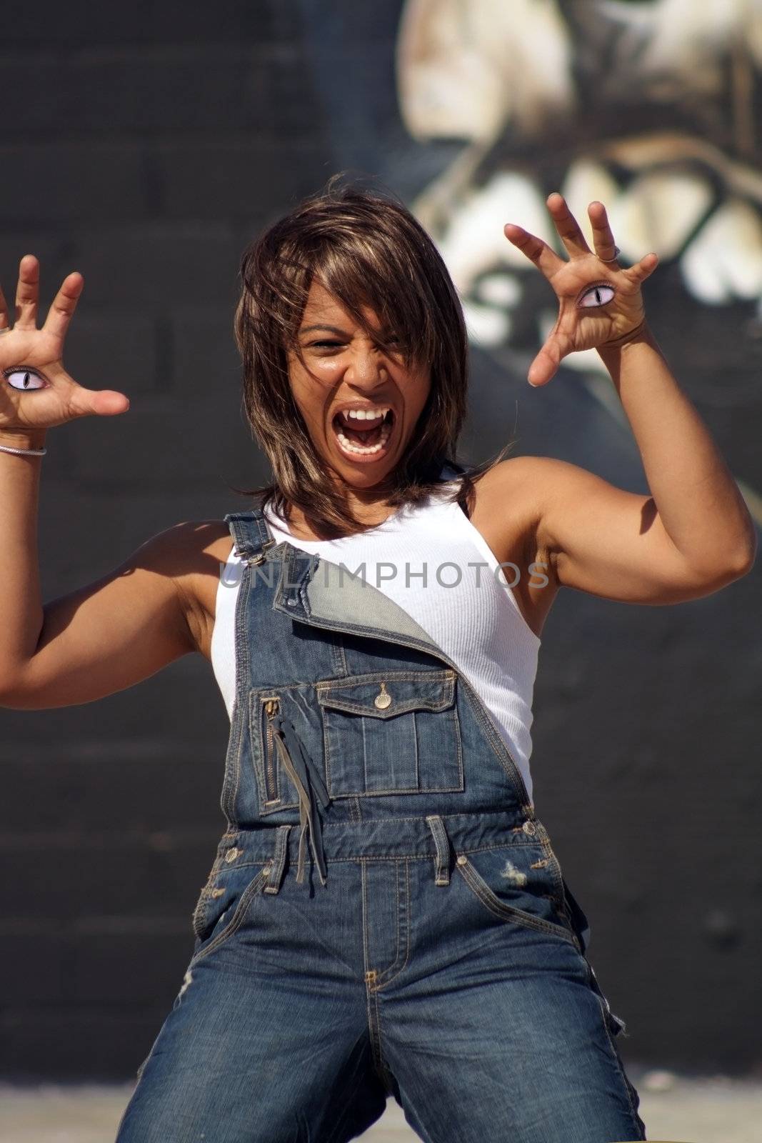 A lovely but scary vampire-ish black woman outdoors, dressed in denim bib overalls with a white tank top, with eyes in the palms of her hands.