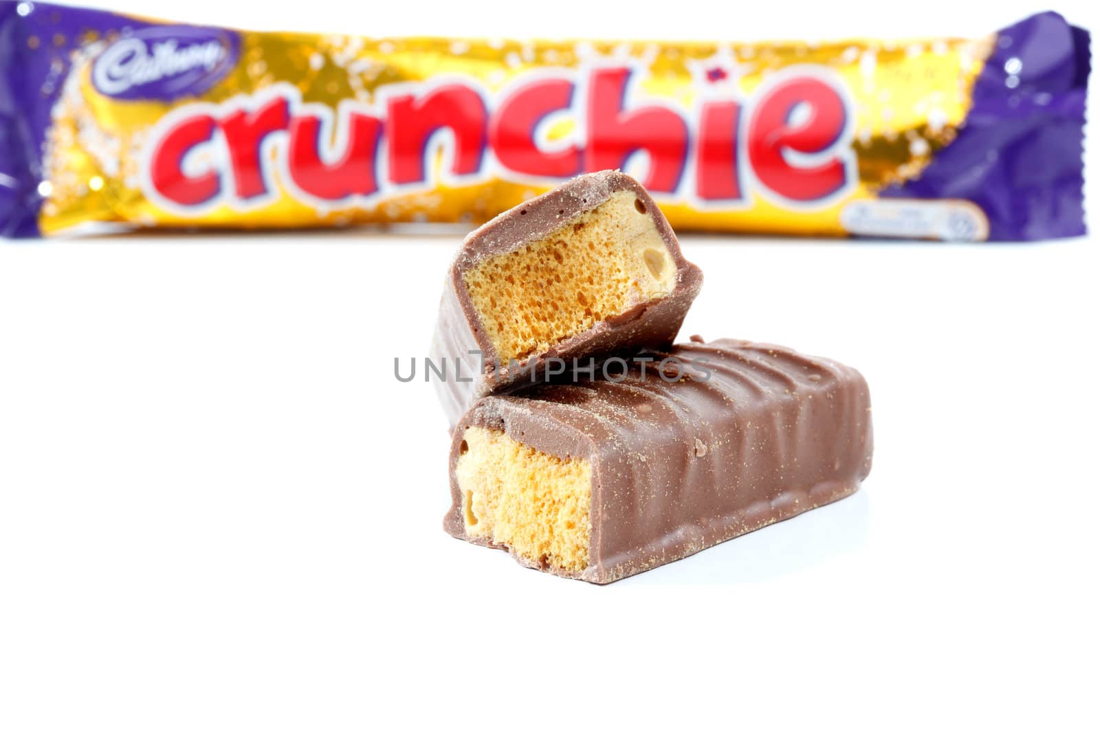 Cadbury Crunchie chocolate coated honeycomb bar. 50g (1015kj) Showing packaging in background and chocolate bar contents in focus in the foreground.  Photographed in studio on a white background.