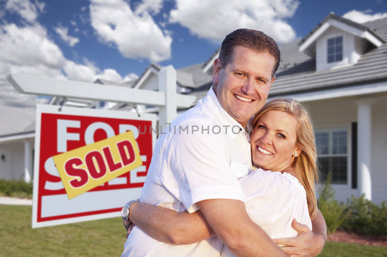 Happy Couple Hugging in Front of Sold Real Estate Sign and House.