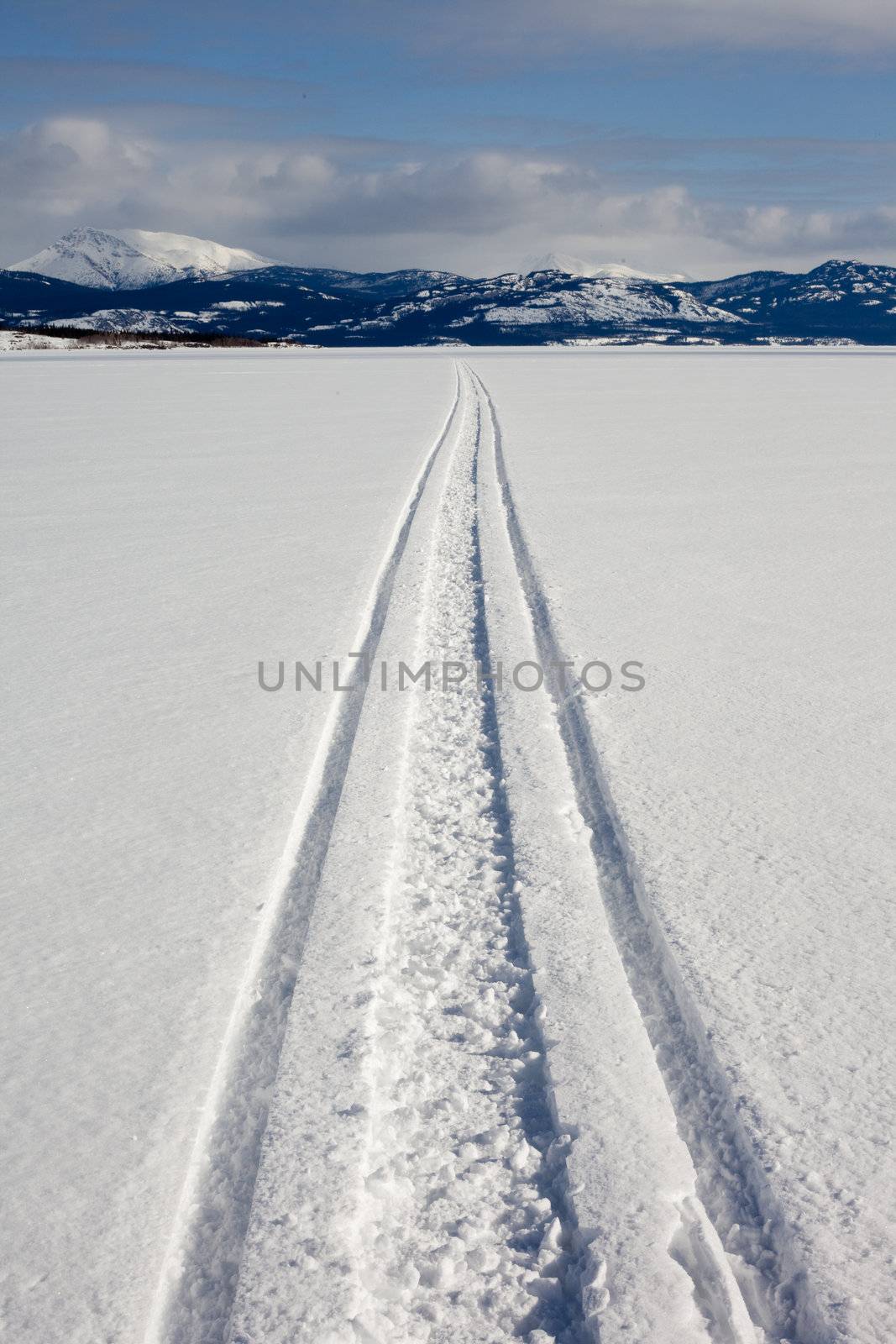 Snowmobile track in snow on surface of frozen lake leading to distant shore on sunny winter day.