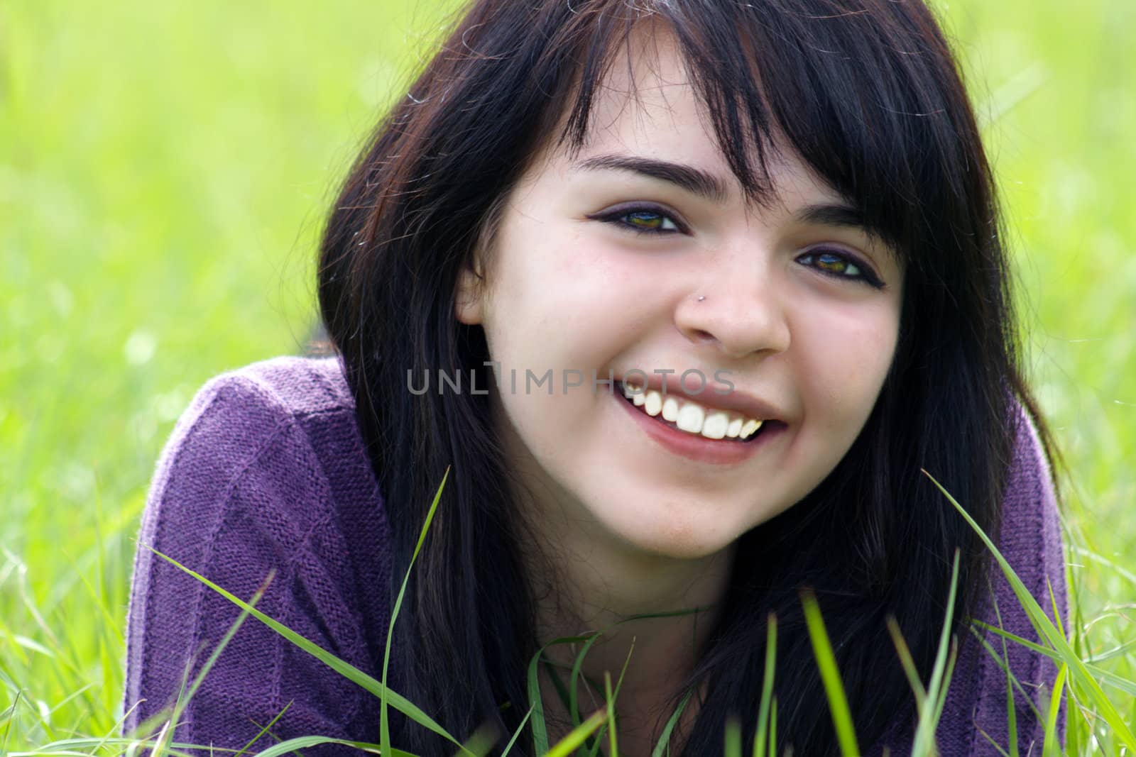 Close-up of a lovely young brunette with a bright, warm smile lying in a field of tall green grass.