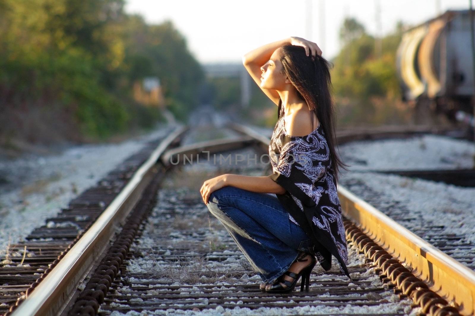 A lovely teenage Latina squats down on a railroad track, facing the sun with her eyes closed.