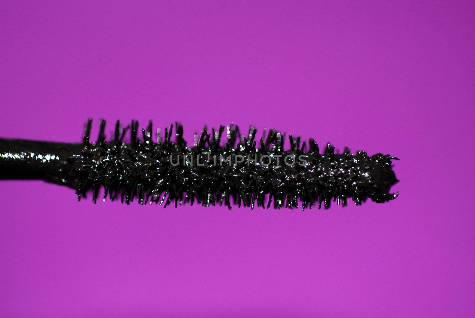 A close-up of the bristles of a mascara wand, with black make-up and a purple background.