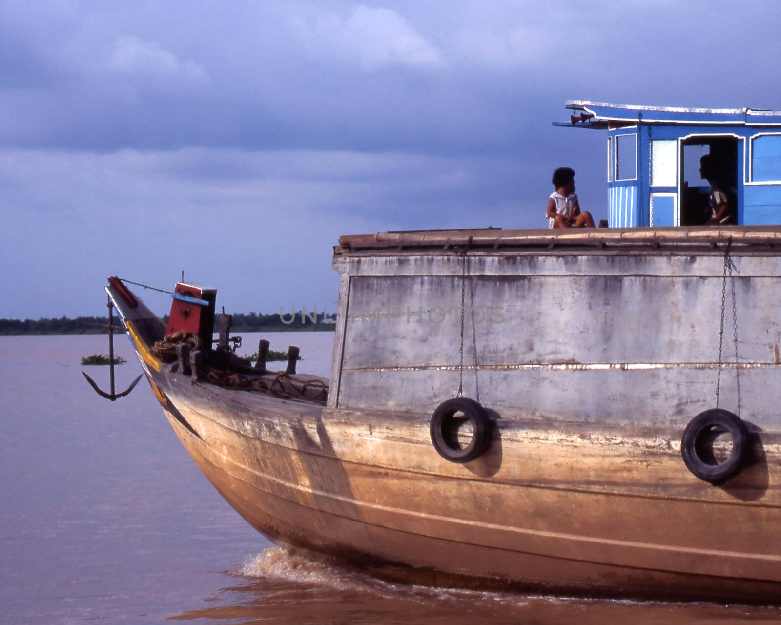 Child sits on roof of wooden house boat. Moving on the Mekong river while in flood.