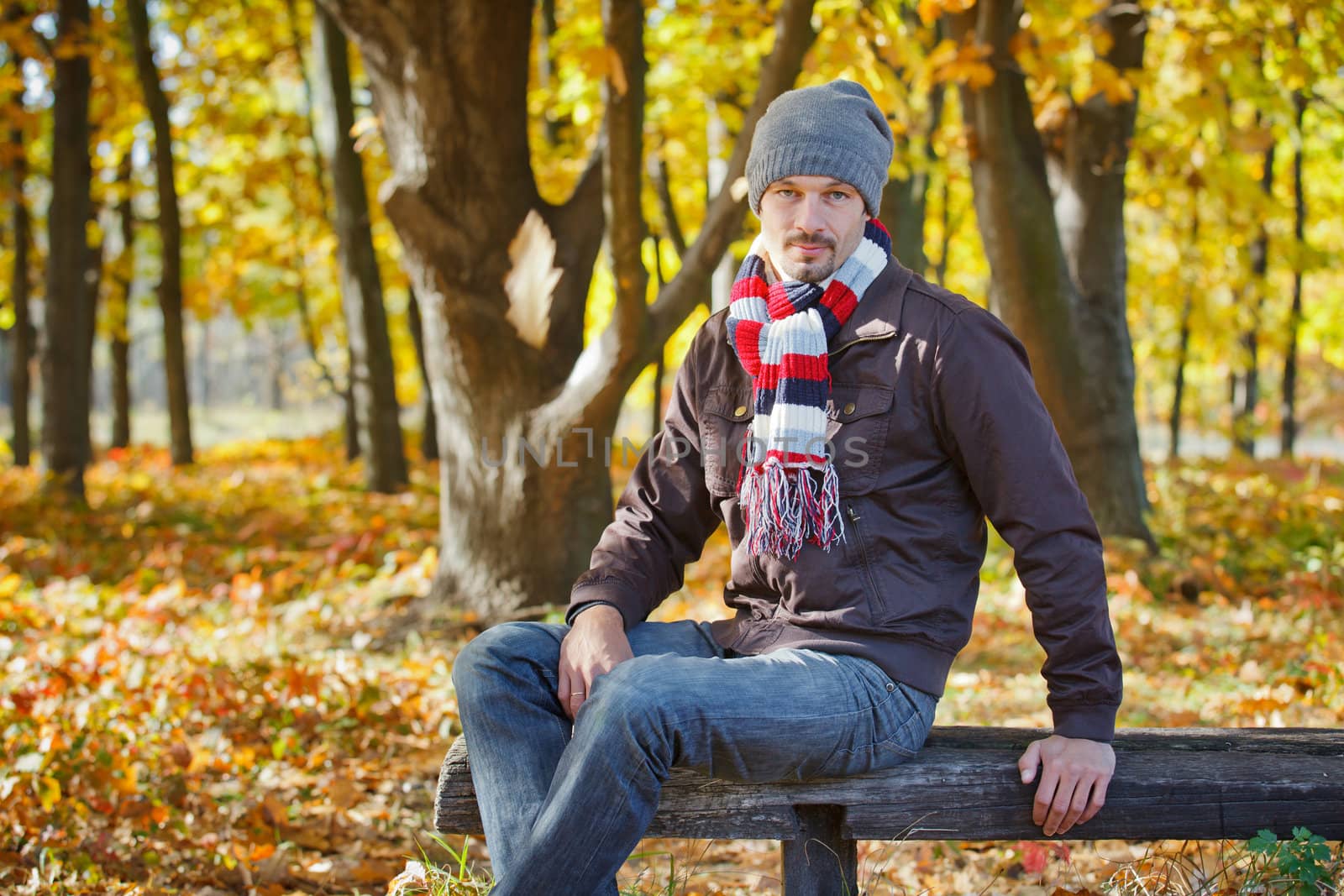 Young attractive man with scarf and hat resting on a bench in autumn park