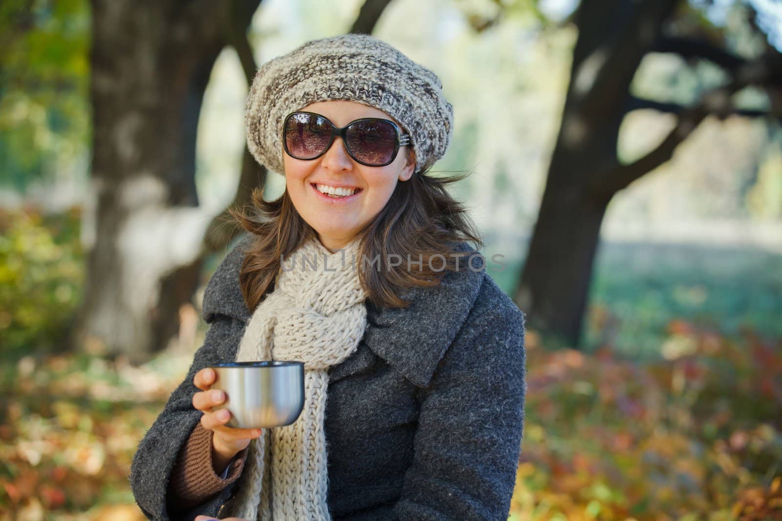 Young pretty woman with glasses and cap drinks tea in autumn park