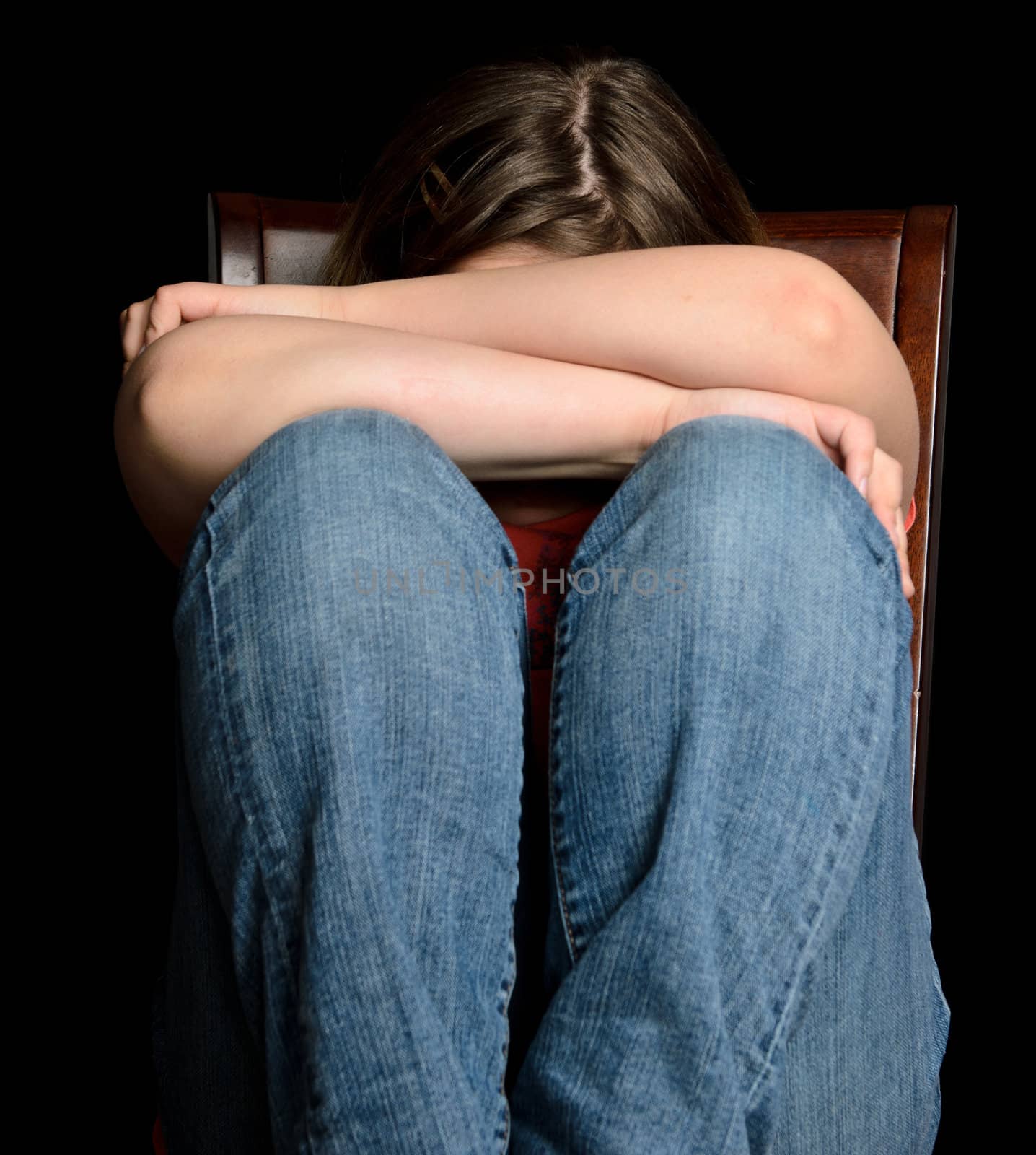 A sad girl holding her head in her arms, while sitting on a chair, isolated on black.