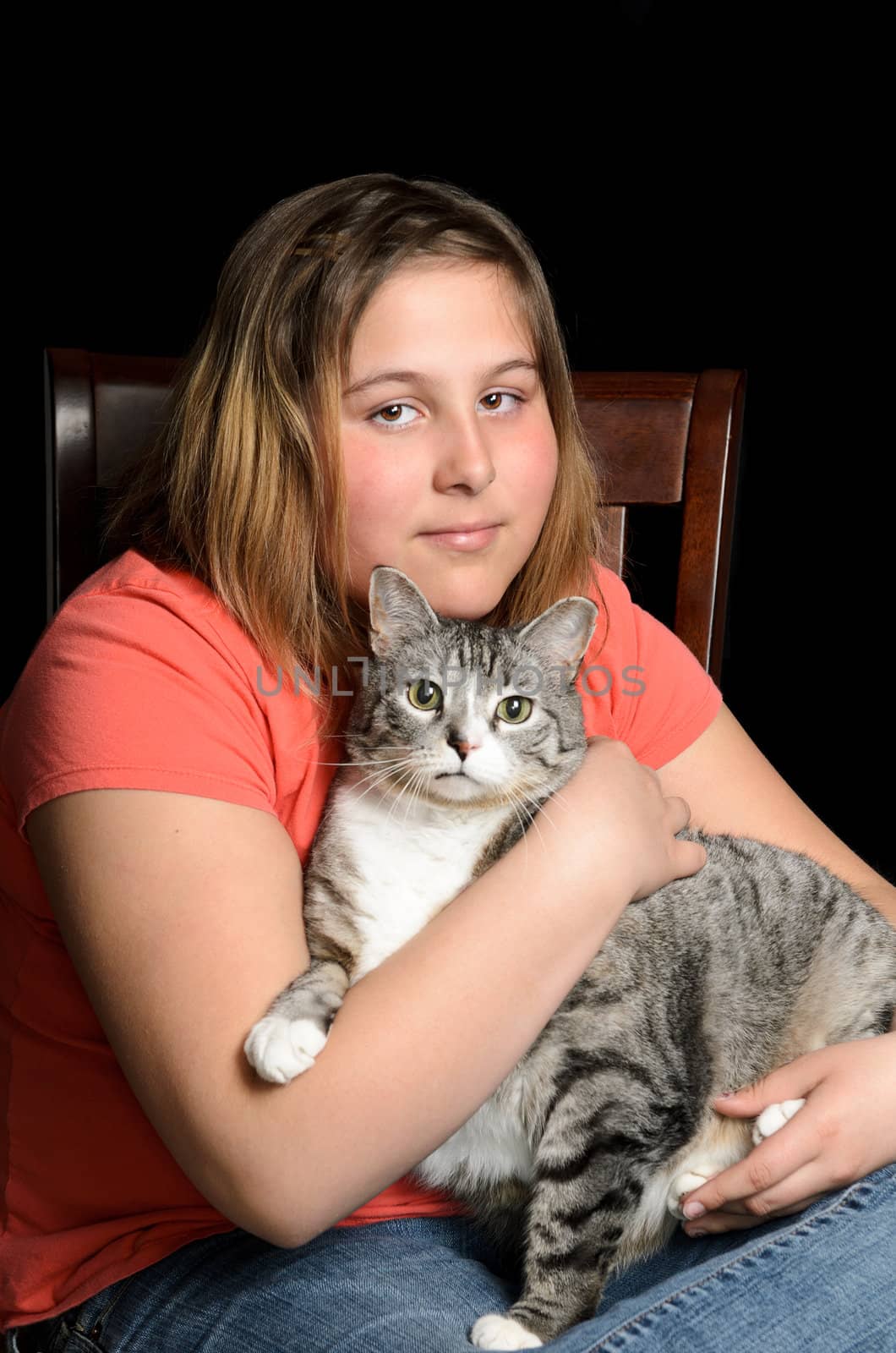 A young girl holding her pet cat, while sitting on a chair, isolated against a black background.