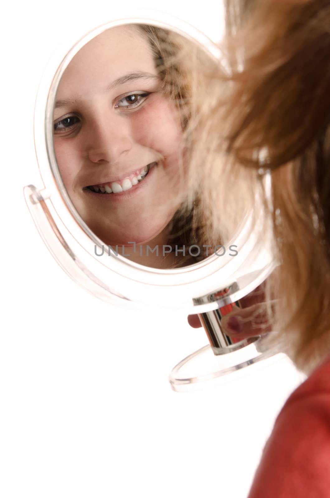 A smiling preteen girl is looking at herself in a small mirror, isolated against a white background.