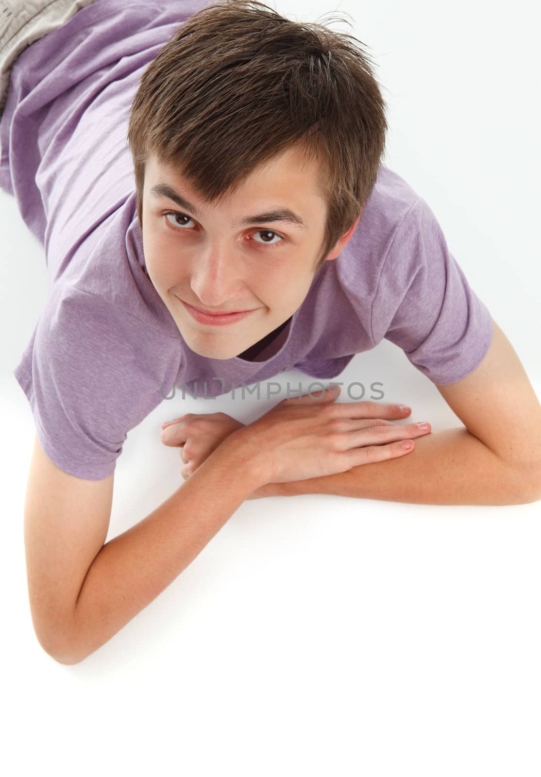 A smiling relaxed boy in a purple t-shirt is resting lying on stomach and looking up.  White background.