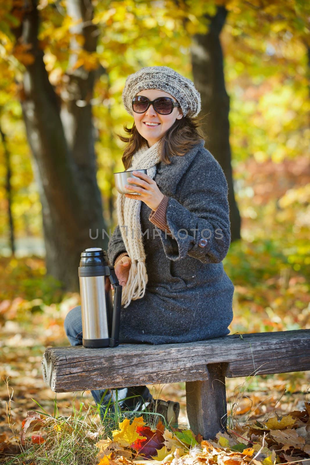 Young pretty woman with glasses and cap drinks tea in autumn park. Vertical view