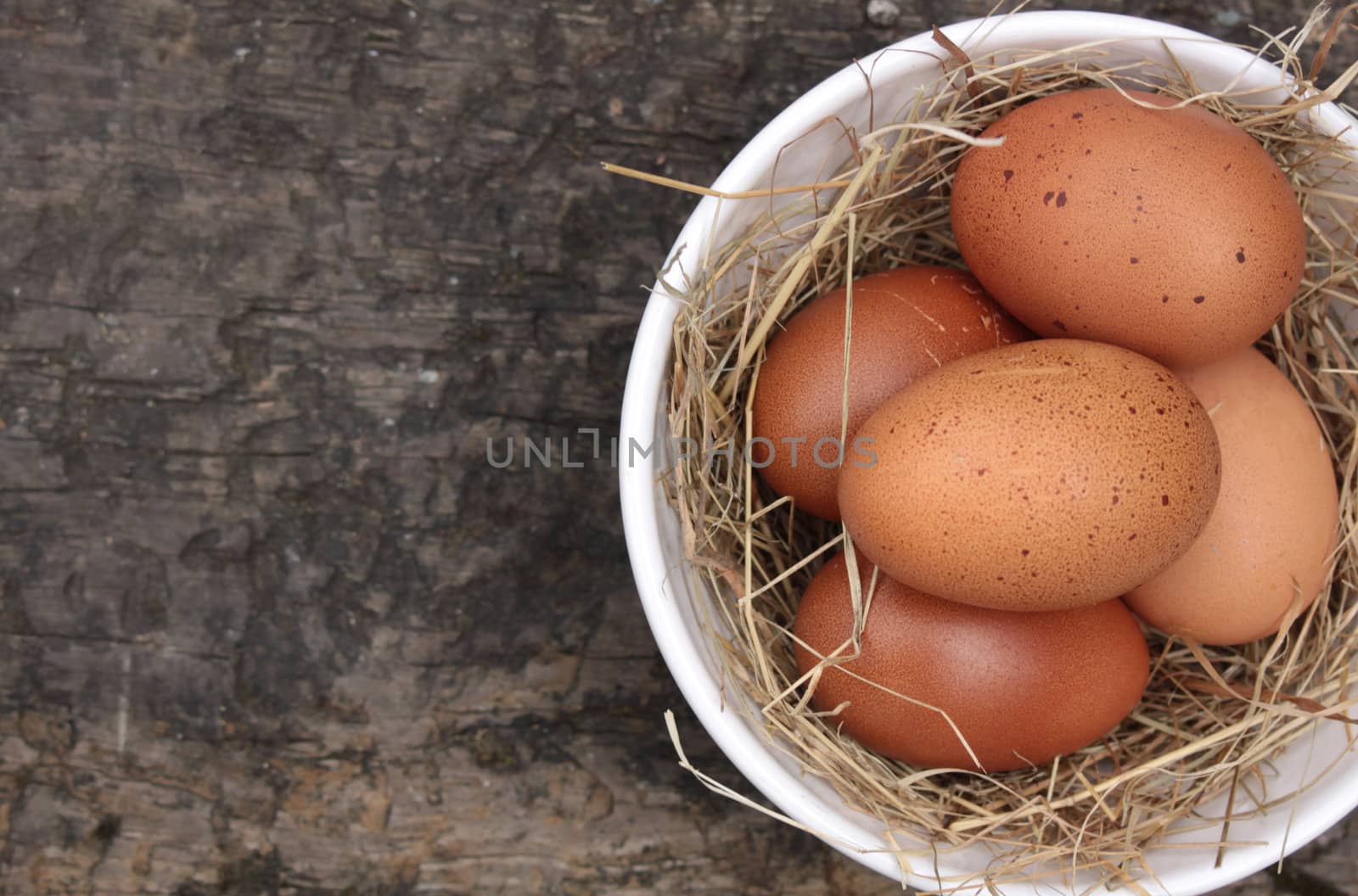 A bowl of freshly laid organic brown eggs, set in a white ceramic bowl with straw. Set on an isolated wooden background. Set on a landscape format with copy space available.