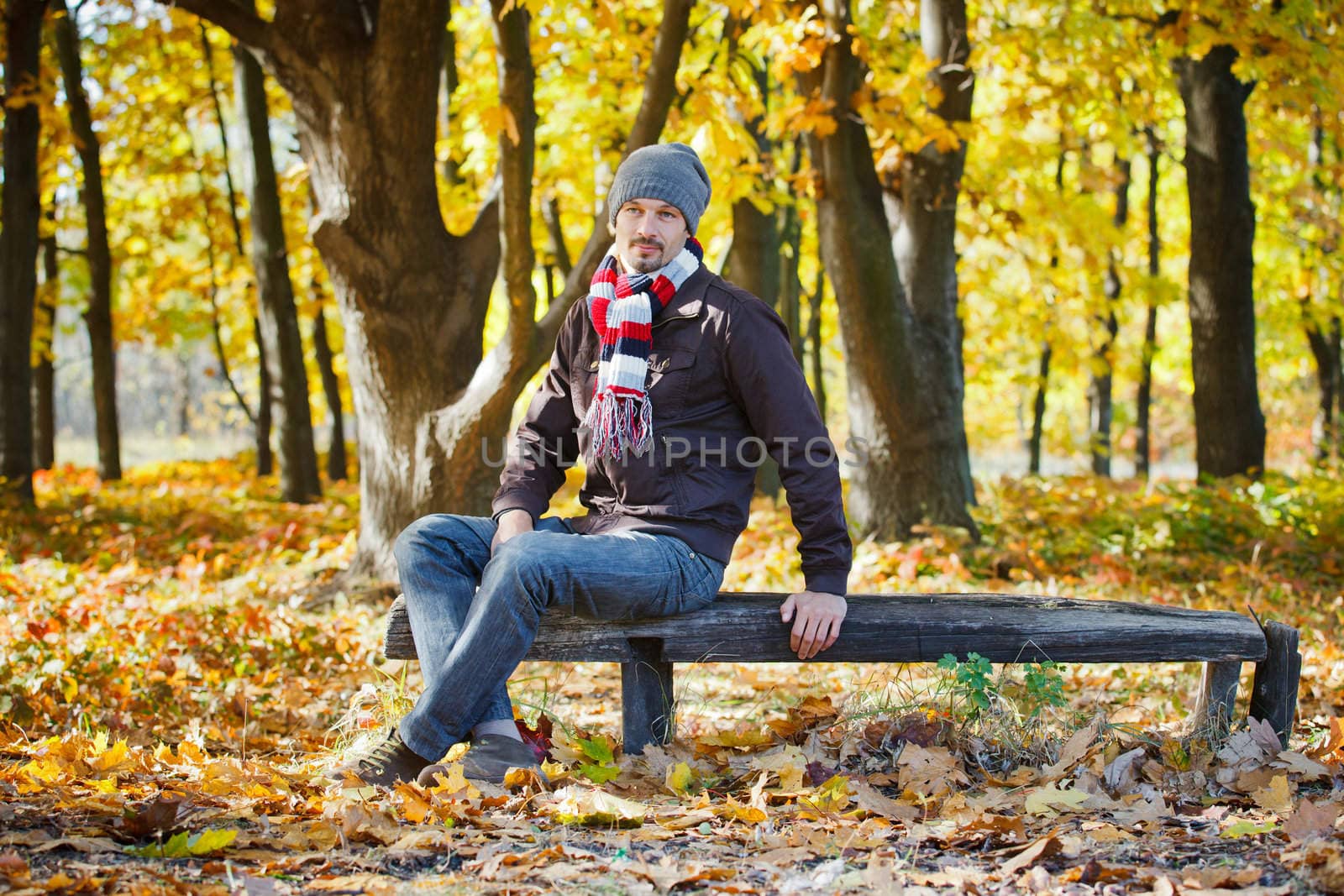 Young attractive man with scarf and hat resting on a bench in autumn park