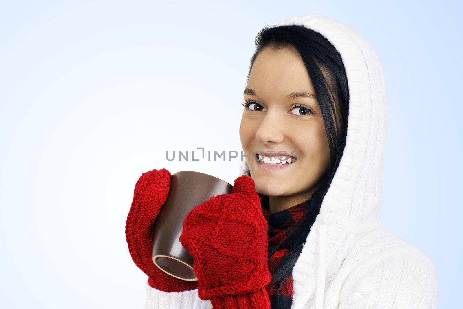 Winter time concept: cute smiling friendly natural young woman with red mittens and white hoodie drinking hot chocolate, coffee or tea in a brown mug over light blue gradient background.