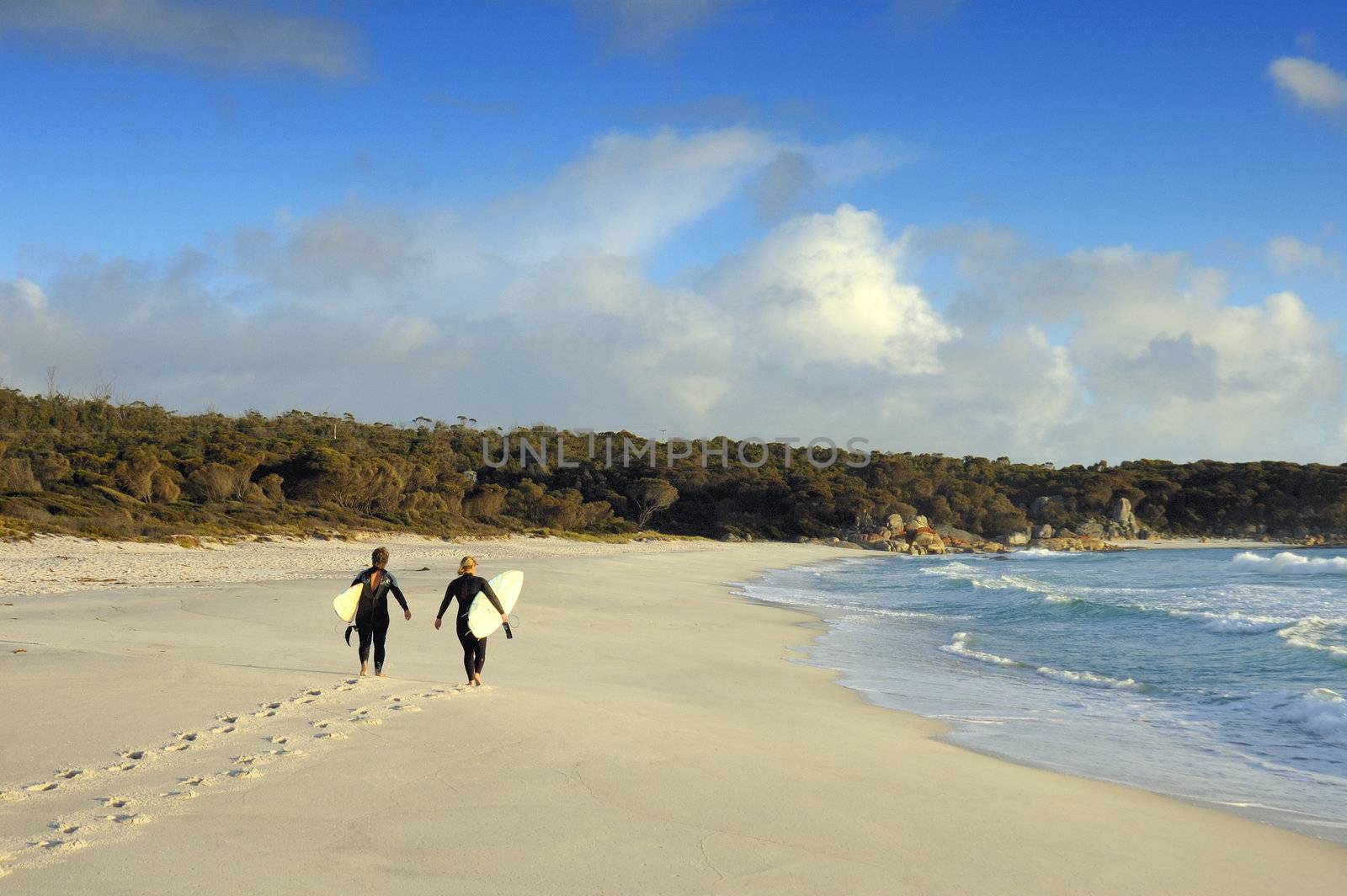 Two women surfers walk along a sandy beach on the Bay of Fires, Tasmania. Photo taken in the early morning.. Space for copy in the sky.