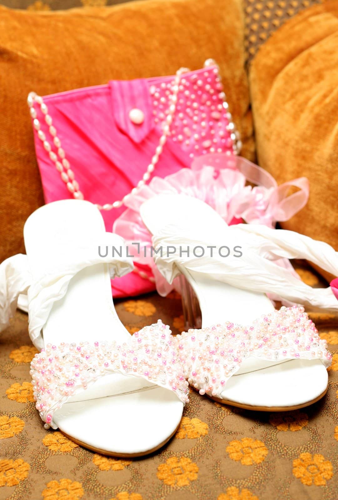 White bridal shoes adorned with pink, white and silver beads with a pink bag and garter in the background lying on brown chair