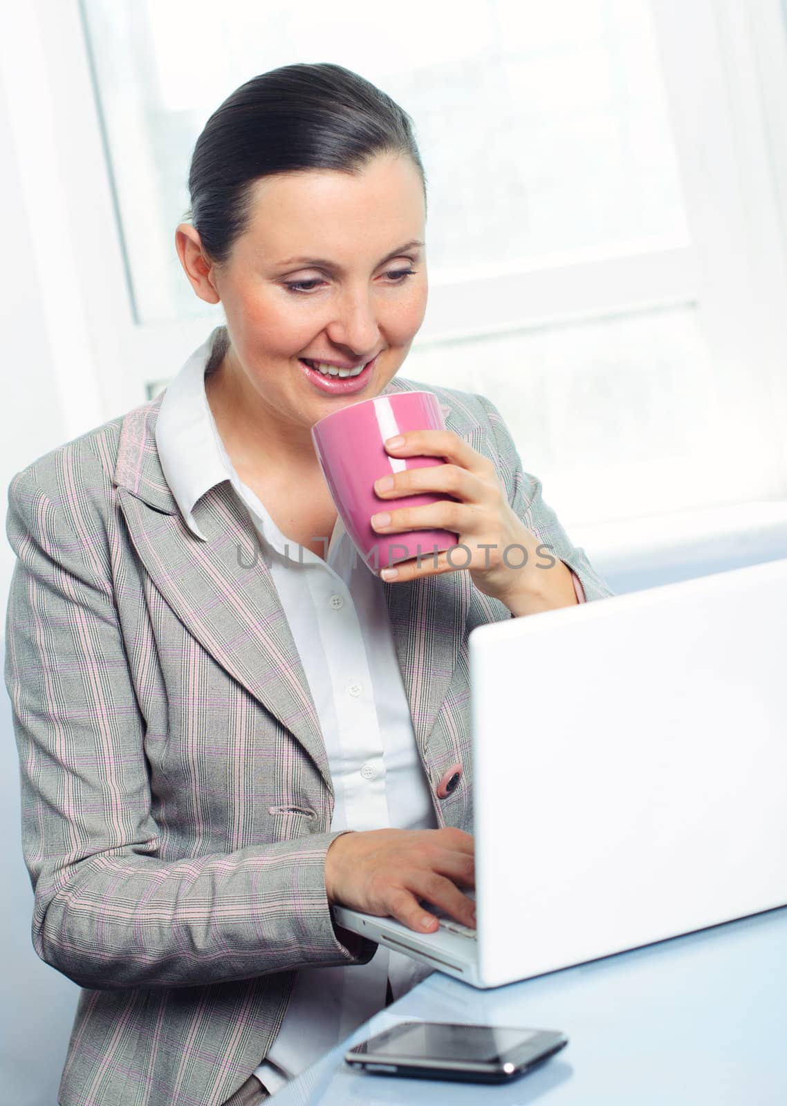 Attractive smiling young business woman with cup using laptop at work desk