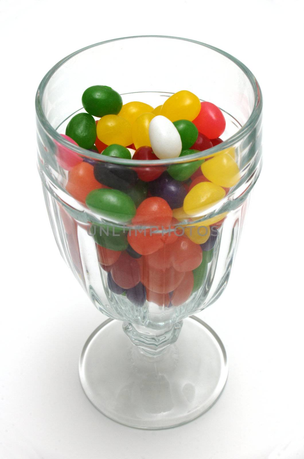A glass sundae cup mostly filled with colorful jelly beans. The view is from slightly above, vertically oriented over white.