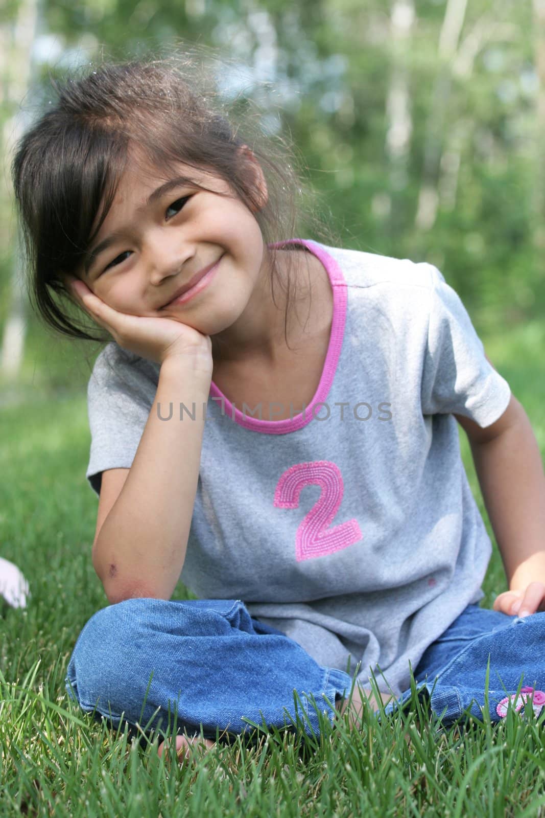 Child sitting on grass with head on hands, smiling. Part asian, scandinavian background.