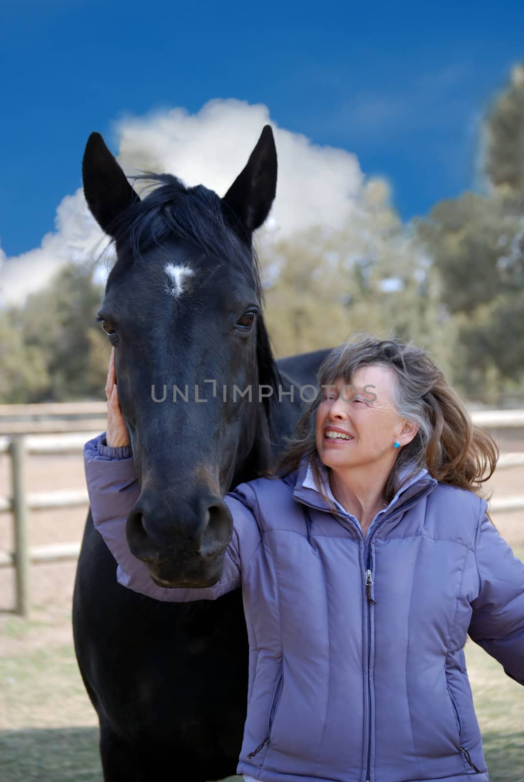 Vertical image of a woman of the Baby Boomer era smiling broadly at her beautiful black horse, against a blue cloudy sky.
