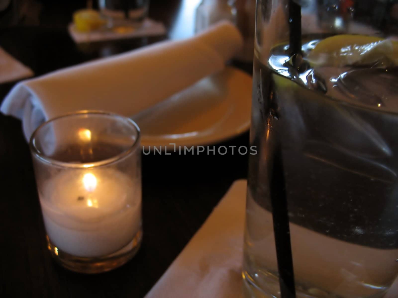 A closeup of a table at a restaurant - waiting to be served.  in the foreground is some water with lemon, and a romantic little candle.