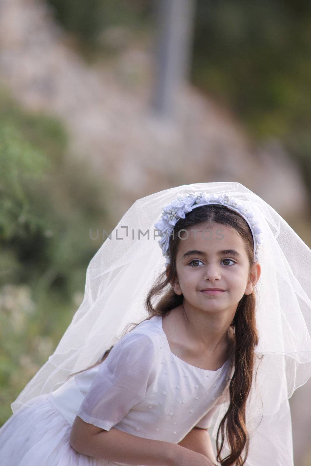 A young girl with long brown haid and big hazel eyes is standing in the countryside dressed in veil and white dress, for her first communion. Vertical photo with a positive energy