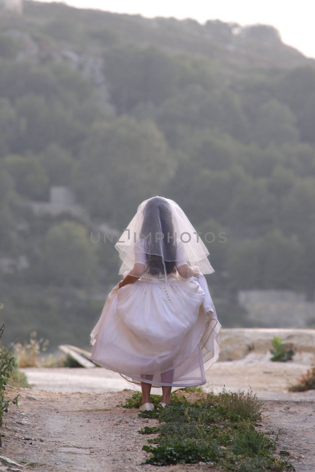 A little girl dressed up as a bride for her Holy Communion is walking away with her back against the camera